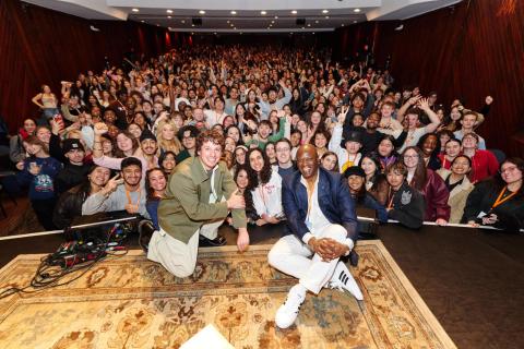 Prince Charles Alexander and Charlie Puth crouch on a stage with a packed audience of students in the background