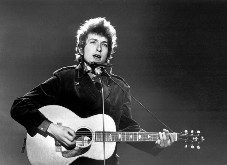 Black and white image of a young Bob Dylan performing on stage with an acoustic guitar and harmonica