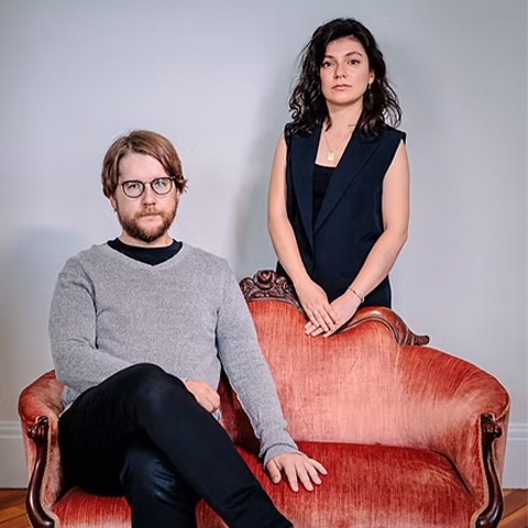 Lilit Hartunian, right,standing in back of a peach-colored couch, wearing a dark shirt and Nicholas Tolle, left, sitting on the couch wearing a grey sweatshirt and black pants.
